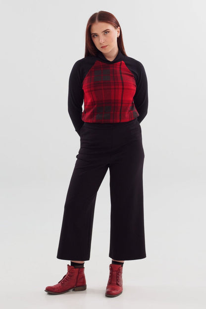 A woman wearing the Nena Sweater by Slak in Red and Black plaid, with contrasting black raglan sleeves, and ribbing at hem, cuffs and cross-over neckline. She is wearing it with black pants and standing in front of a white background 