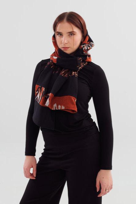 A woman wearing the Slak A24 Scarf in Black and Caramel, a long open-ended scarf made contrasting strips of leftover fabric from the current collection. She is wearing it with a black top and black pants and is standing in front of a white background 