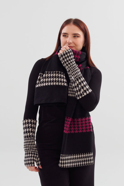 A woman wearing the Slak A24 Scarf in Black and White and Pink, a long open-ended scarf made contrasting strips of leftover fabric from the current collection. She is wearing it with a black top and black pants and is standing in front of a white background 