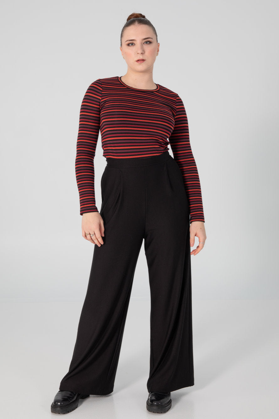 A woman wearing the A25 Sweater by Slak in Colourful Stripe, a long sleeved top with a round neck and a slim fit. She is wearing it with black skirt pants and standing in front of a white background. 