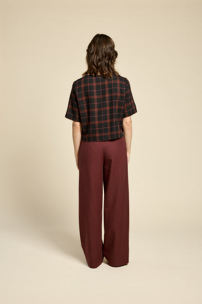 Back view of a woman wearing the Soul Love Top by Cokluch in Burgundy Plaid, with a round neck, loose fit, and elbow length sleeves. She is wearing it with with burgundy pants and standing against a beige background.