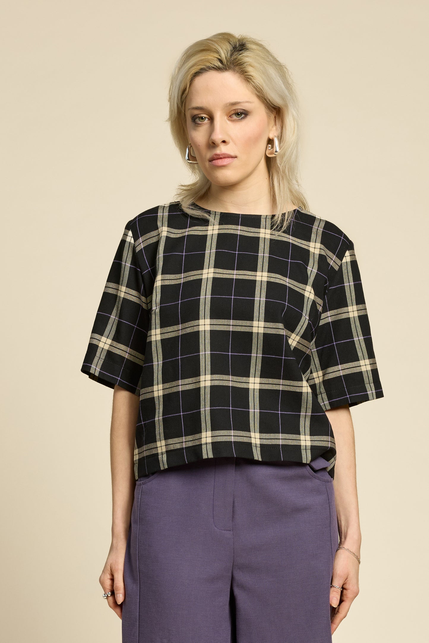 A woman wearing the Soul Love Top by Cokluch in Black and beige plaid, with a round neck, loose fit, and elbow length sleeves. She is wearing it with with purple pants and standing against a beige background.