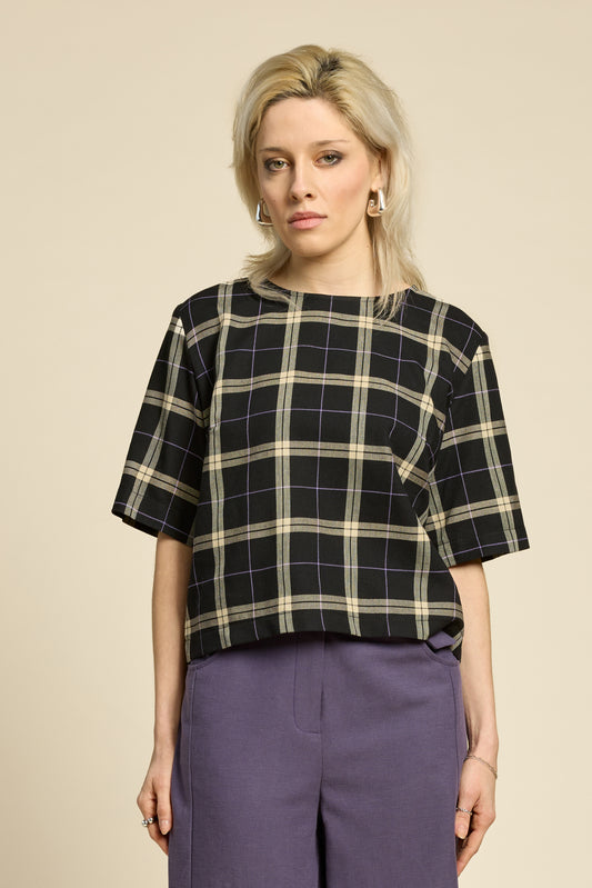 A woman wearing the Soul Love Top by Cokluch in Black and beige plaid, with a round neck, loose fit, and elbow length sleeves. She is wearing it with with purple pants and standing against a beige background.