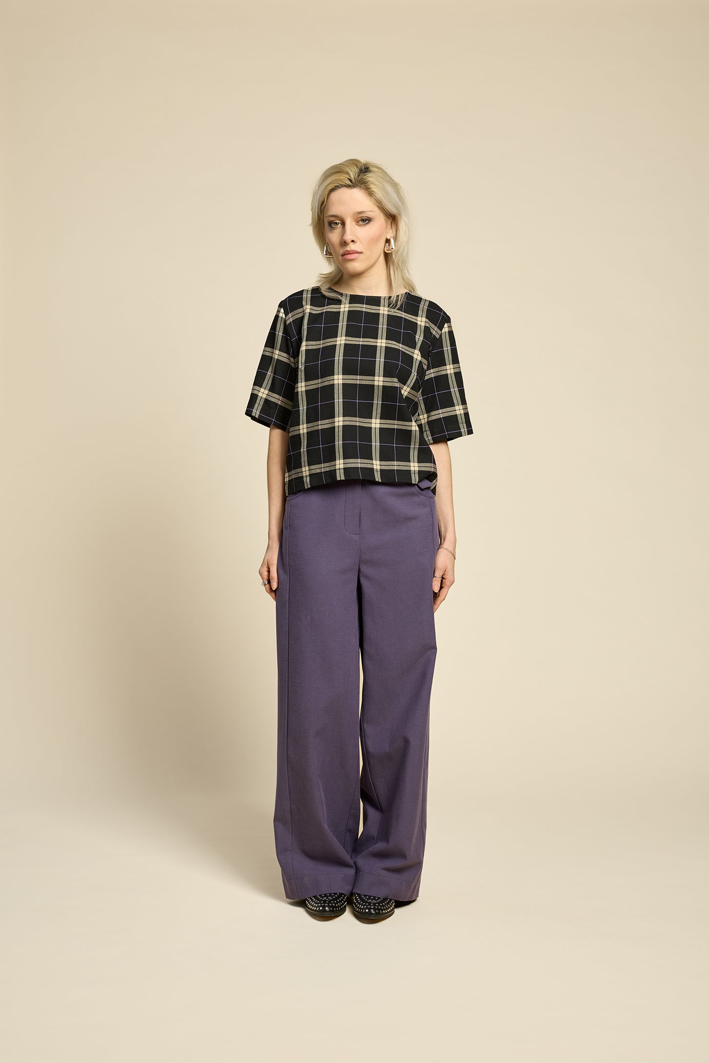 A woman wearing the Soul Love Top by Cokluch in Black and beige plaid, with a round neck, loose fit, and elbow length sleeves. She is wearing it with with purple pants and standing against a beige background.