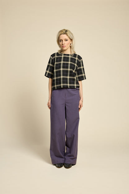 A woman wearing the Soul Love Top by Cokluch in Black and beige plaid, with a round neck, loose fit, and elbow length sleeves. She is wearing it with with purple pants and standing against a beige background.