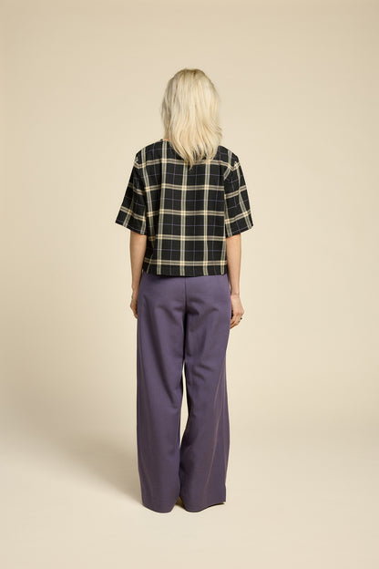 Back view of A woman wearing the Soul Love Top by Cokluch in Black and beige plaid, with a round neck, loose fit, and elbow length sleeves. She is wearing it with with purple pants and standing against a beige background.
