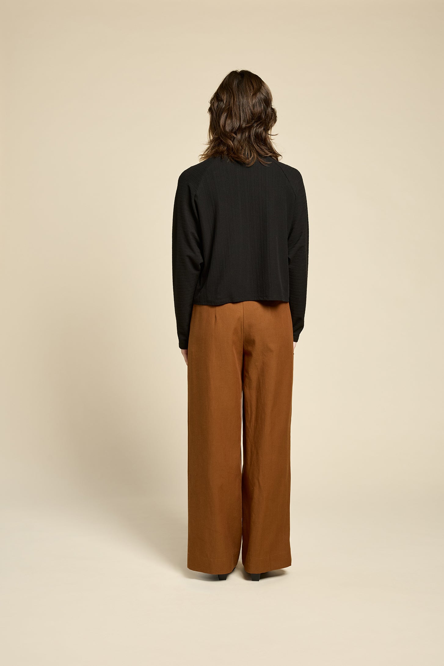 Back view of a woman wearing the Souvenir Top by Cokluch in Sand, with a twisted turtleneck, ribbed fabric with a vertical band on the front. She's wearing it with burgundy pants and standing in front of a beige background. 