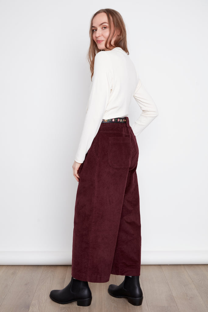 Back view of a woman wearing the Melody Mockneck Top by Julie in Natural, a ribbed knit top with a short cross-over collar and long sleeves. She is wearing it with the Isadora Pants in Burgundy corduroy against a white background.