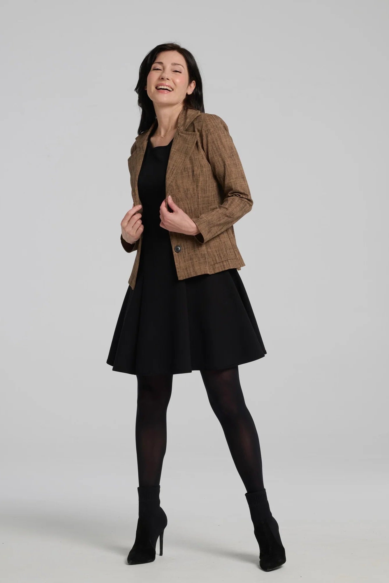 A woman wearing the Toscana Dress by Kollontai in Black, a sleeveless piece with a round neck, princess seams, and fit and flare shape with a full skirt that hits above the knee. She is wearing it with a brown blazer and standing against a grey background.