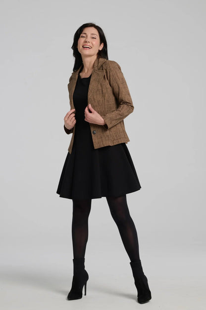 A woman wearing the Toscana Dress by Kollontai in Black, a sleeveless piece with a round neck, princess seams, and fit and flare shape with a full skirt that hits above the knee. She is wearing it with a brown blazer and standing against a grey background.