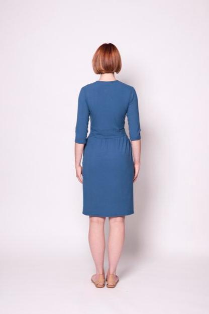 Back view of a woman wearing the Camille Dress by Tangente in Blue, featuring a cross-over neckline, narrow waistband, knee length skirt, 3/4 sleeves, and side pockets, in ribbed bamboo knit. She is standing in front of white background 
