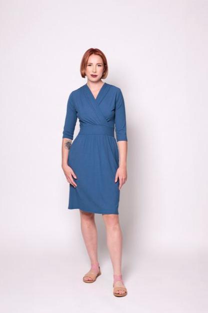 A woman wearing the Camille Dress by Tangente in Blue, featuring a cross-over neckline, narrow waistband, knee length skirt, 3/4 sleeves, and side pockets, in ribbed bamboo knit. She is standing in front of white background 