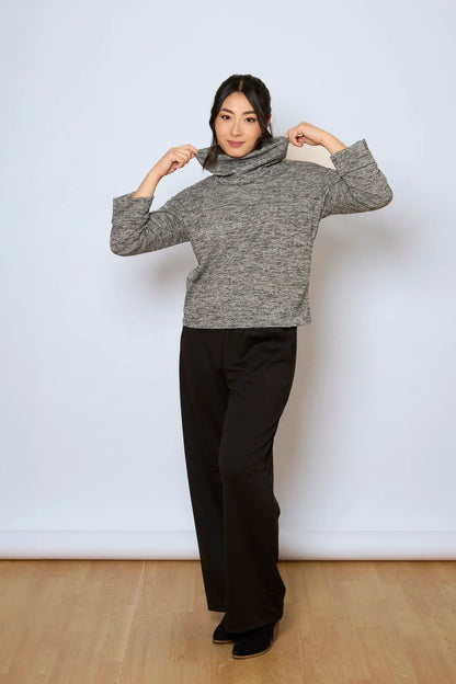 A woman wearing the Jill Pants by Tangente in Black, wide-legged ankle length pants with large pockets. She is wearing them with a grey sweater against a white background.