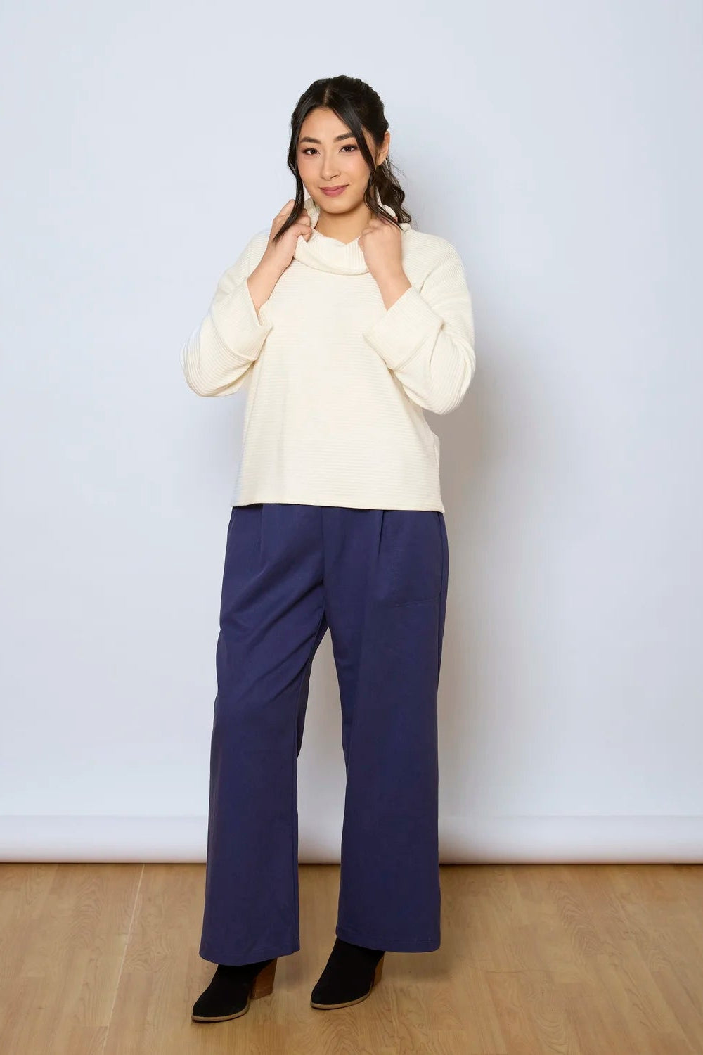 A woman wearing the Jill Pants by Tangente in Denim, wide-legged ankle length pants with large pockets. She is wearing them with a white sweater against a white background.