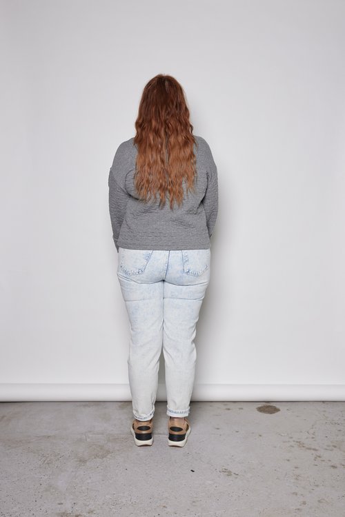 Back view of a woman wearing the Sable Top by Tangente in Grey, a sweatshirt in quilted fabric with a cowl neck and cuffed sleeves and a slightly cropped and boxy fit. She is wearing it with jeans and standing in front of a white background. 