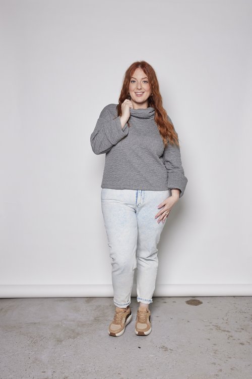 A woman wearing the Sable Top by Tangente in Grey, a sweatshirt in quilted fabric with a cowl neck and cuffed sleeves and a slightly cropped and boxy fit. She is wearing it with jeans and standing in front of a white background. 