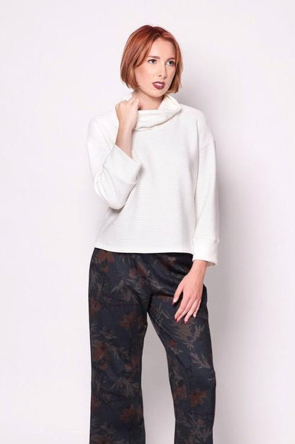 A woman wearing the Sidney Top by Tangente in Off-white, a thick ribbed knit sweater with a cowl neck, long cuffed sleeves, and a slightly boxy fit. She's wearing it with floral pants and standing in front of a white background. 