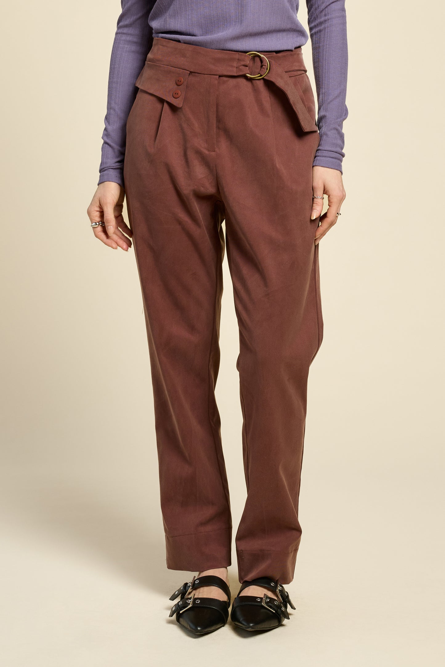Waist-down view of a woman wearing the Fool Pants by Cokluch in Cappucino, featuring an attached belt with D-rings, a flap and button detail at the waist, and tapered ankle-length legs. She is wearing them with a purple top and standing in front of a beige background