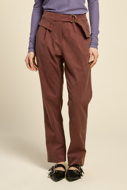 Waist-down view of a woman wearing the Fool Pants by Cokluch in Cappucino, featuring an attached belt with D-rings, a flap and button detail at the waist, and tapered ankle-length legs. She is wearing them with a purple top and standing in front of a beige background