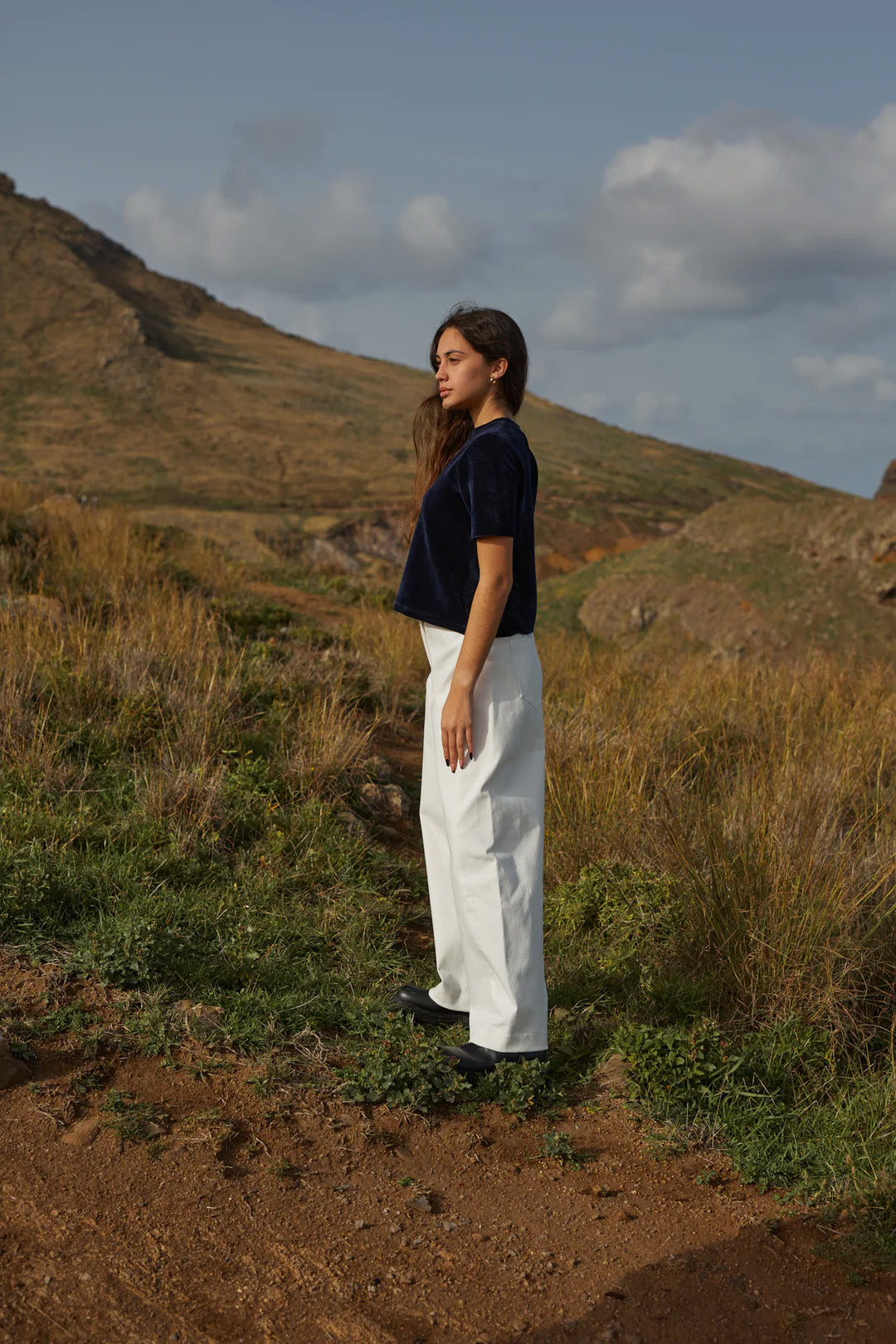 Side view of a woman wearing the Tofino Top by MAS in Indigo, a slightly cropped tee in velvet rib. She is wearing it with white pants and is standing on a hill. 