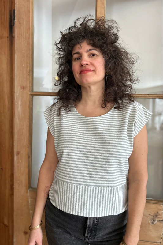 A woman wearing the Tulipe Top by Kazak in Sage, a short top with a round neck, short extended sleeves, horizontal stripes across the chest and a band of vertical stripes across the bottom, with a rounded hem and side slits. She is wearing it with black jeans and standing in front of a wooden door. 