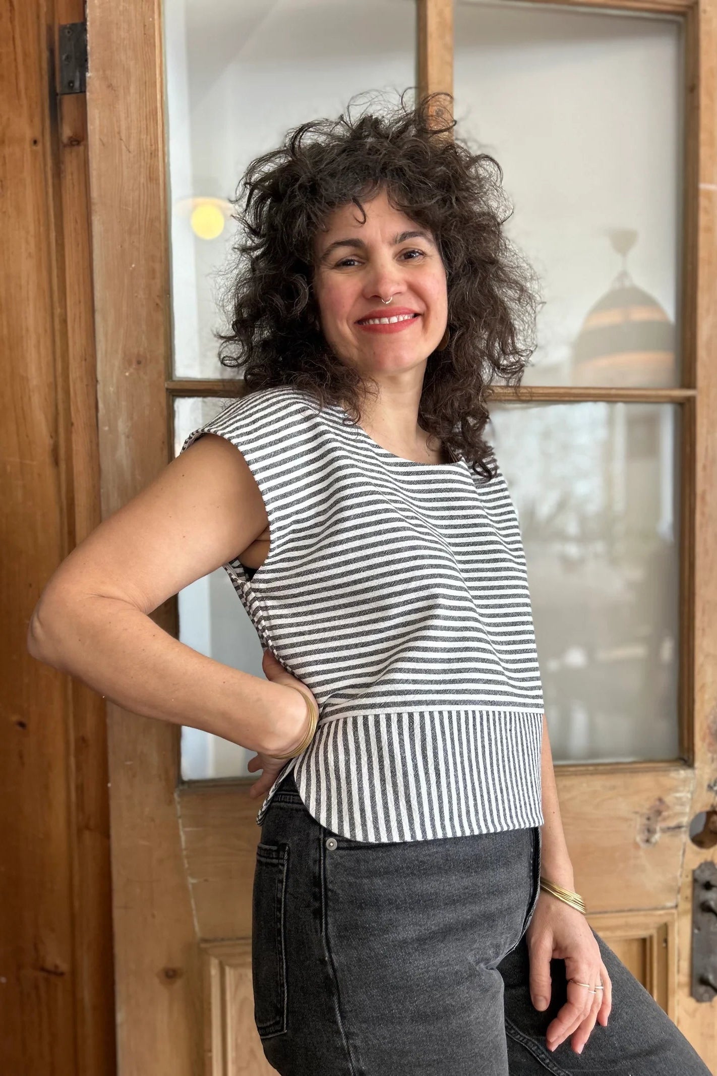 A woman wearing the Tulipe Top by Kazak in Black, a short top with a round neck, short extended sleeves, horizontal stripes across the chest and a band of vertical stripes across the bottom, with a rounded hem and side slits. She is wearing it with black jeans and standing in front of a wooden door. 