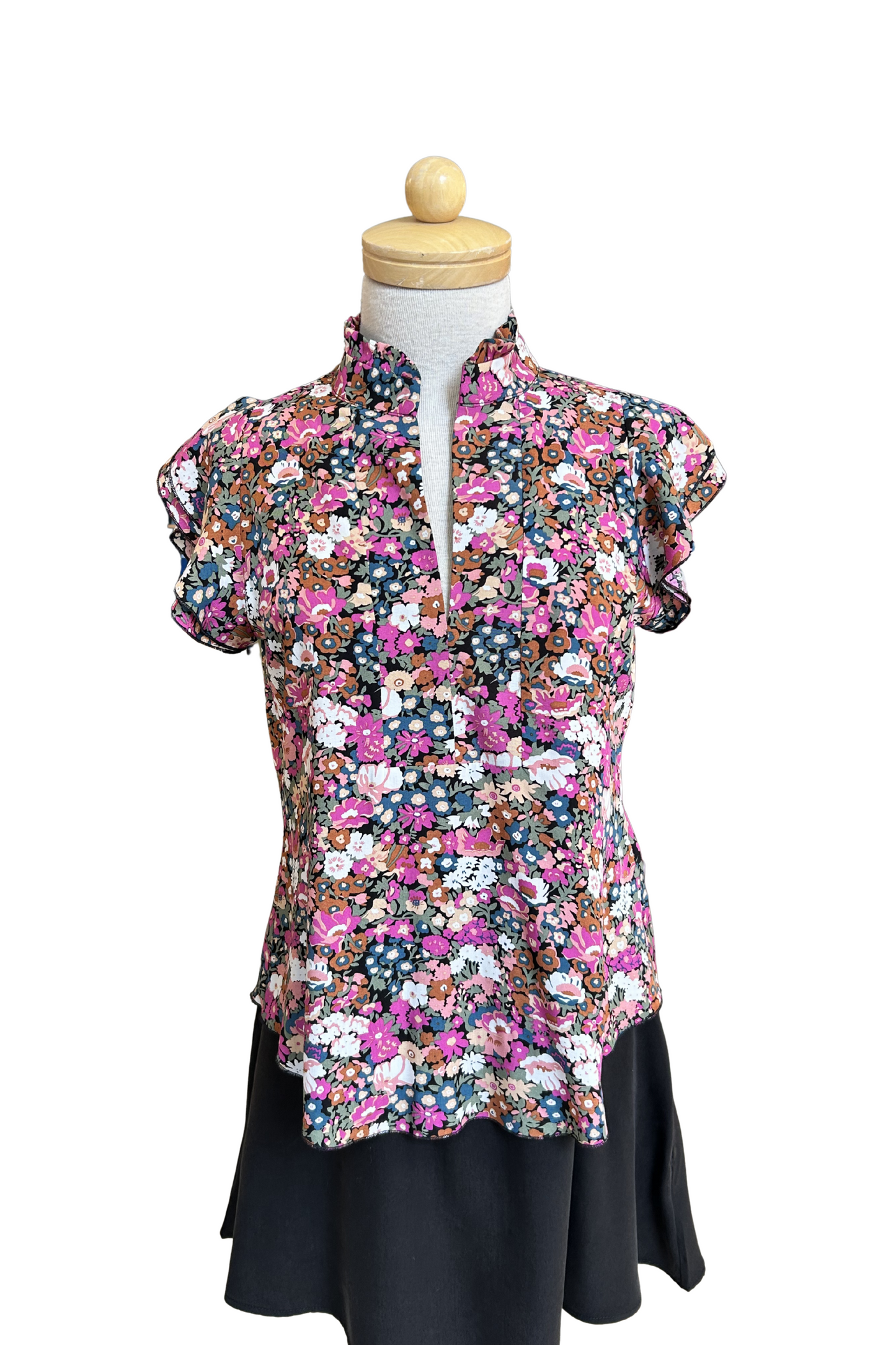 The Petal Blouse by Dotty in Pink/Blue floral print is shown on a mannequin against a white background. It has short floaty sleeves, a split V-neck, a ruffled collar, and a rounded hi-low hemline. 