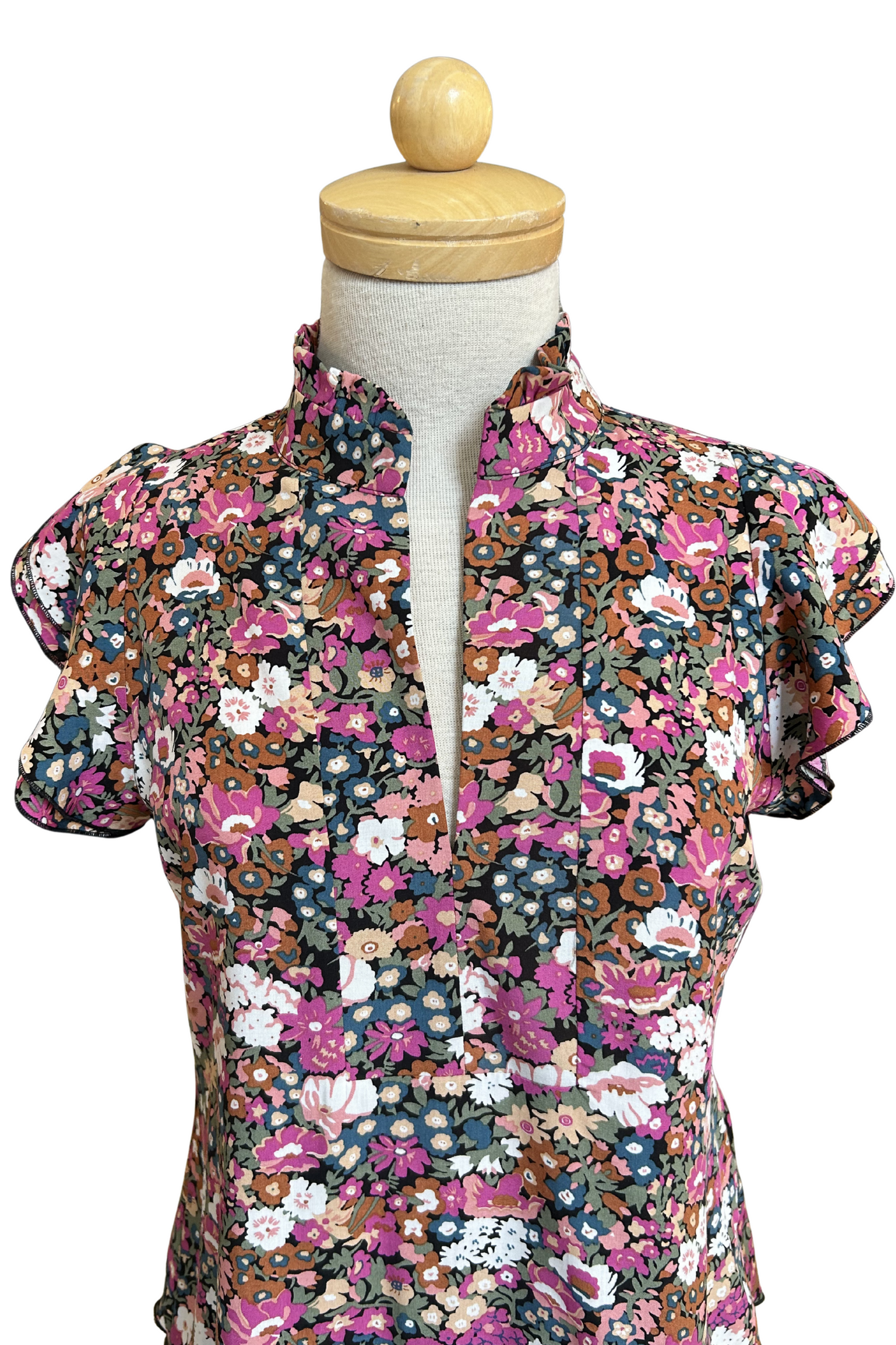 The Petal Blouse by Dotty in Pink/Blue floral print is shown on a mannequin against a white background. It has short floaty sleeves, a split V-neck, a ruffled collar, and a rounded hi-low hemline. 