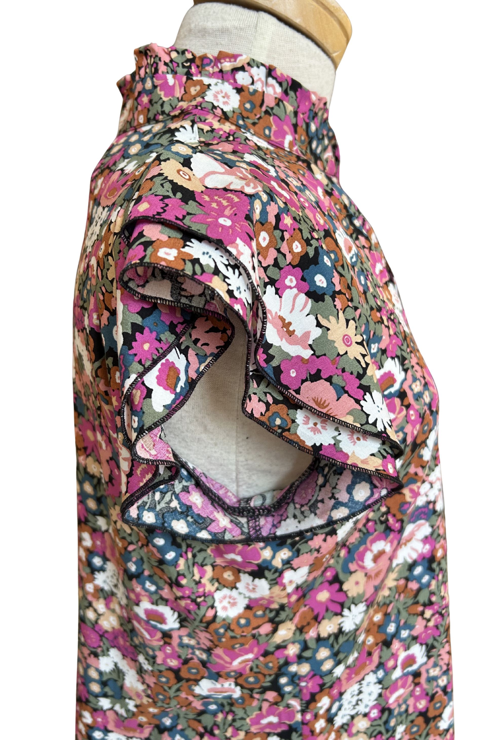 A side view of the Petal Blouse by Dotty in Pink/Blue floral print is shown on a mannequin against a white background. It has short floaty sleeves, a split V-neck, a ruffled collar, and a rounded hi-low hemline. 