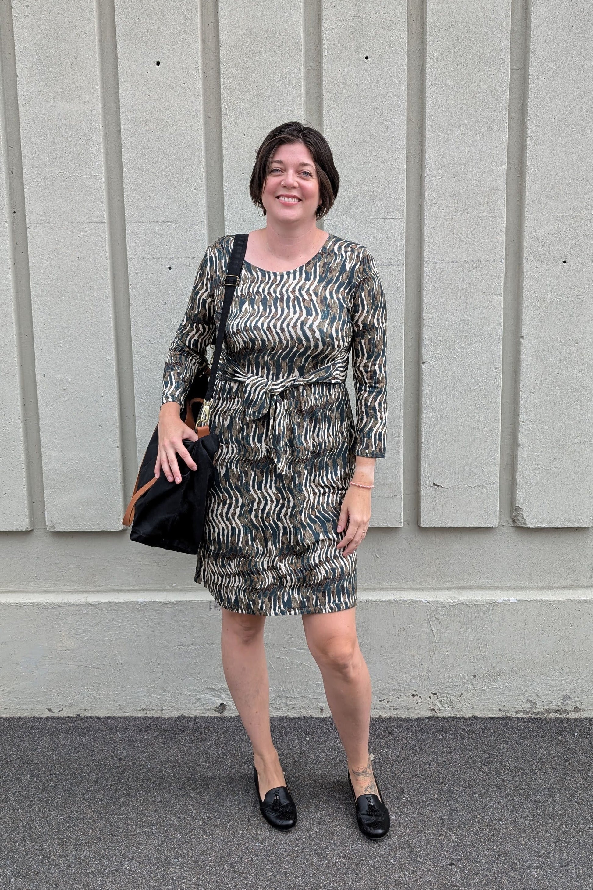 A woman wearing the Gwen Dress by Tangente in Green print, standing outside in front of a white wall
