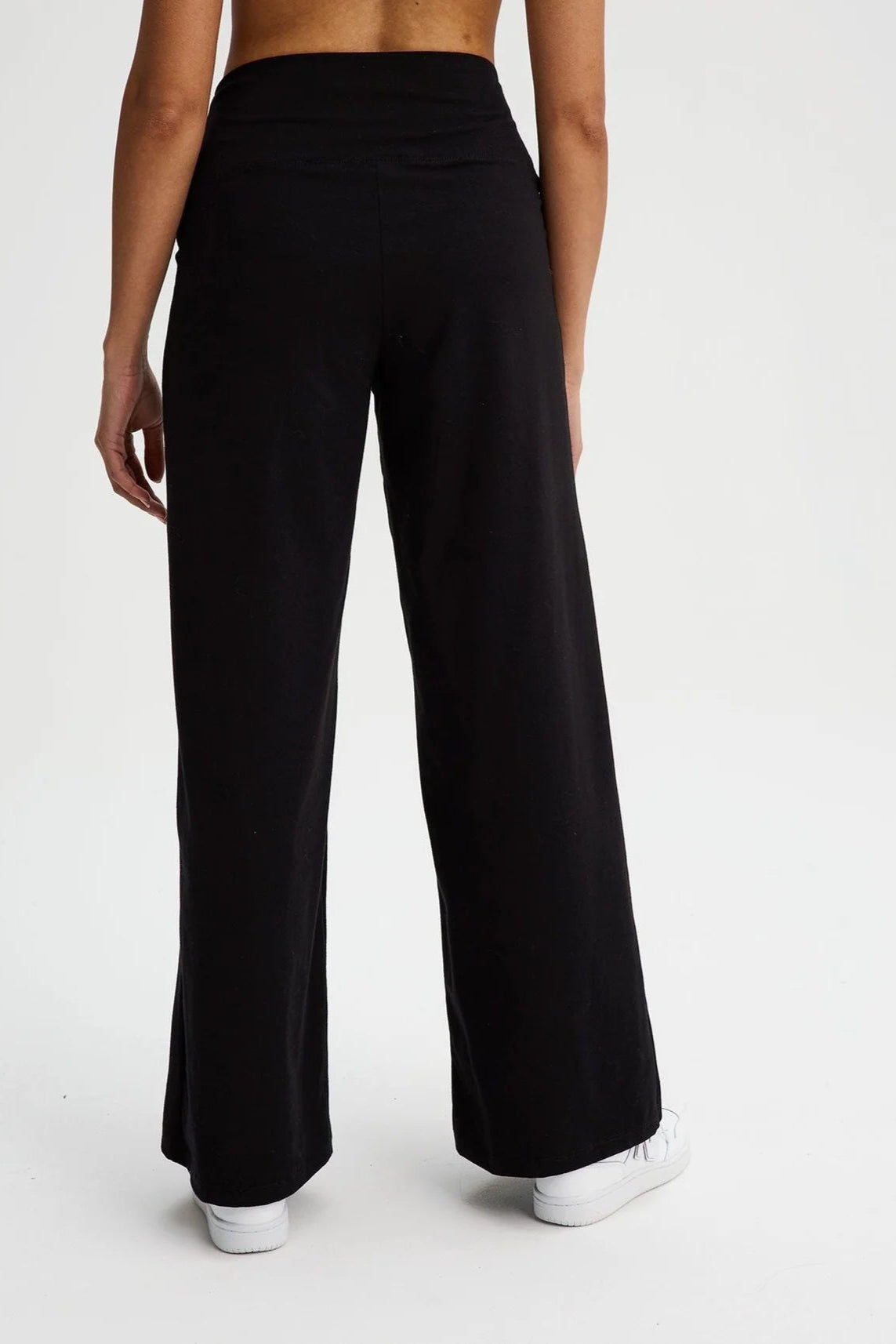 Back waist down view of a woman wearing the Villeray Palazzo Pants by Message Factory in Black, wide legged organic cotton pants with a pull-on elastic waistband with a cross-over detail. She is standing in front of a white background. 