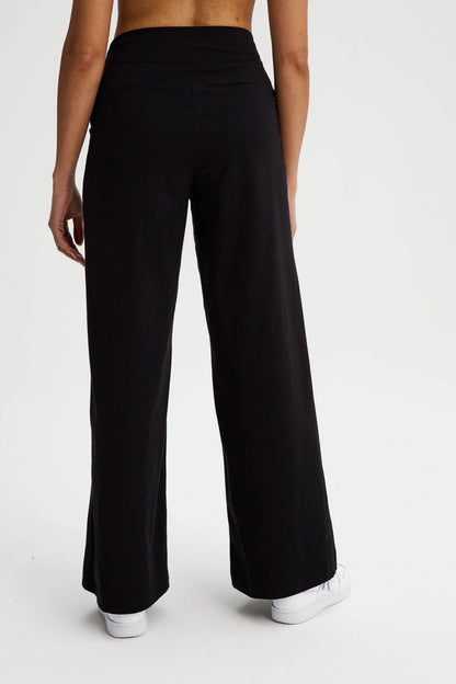 Back waist down view of a woman wearing the Villeray Palazzo Pants by Message Factory in Black, wide legged organic cotton pants with a pull-on elastic waistband with a cross-over detail. She is standing in front of a white background. 