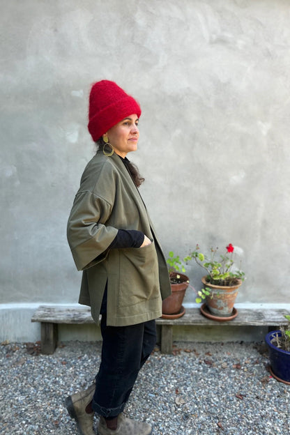 Side view of a woman wearing the Cydonia Cardigan by Kazak in Olive, a hip-length open cardigan with 3/4 kimono sleeves and large patch pockets. She is wearing it with black pants and a black top and standing outside. 