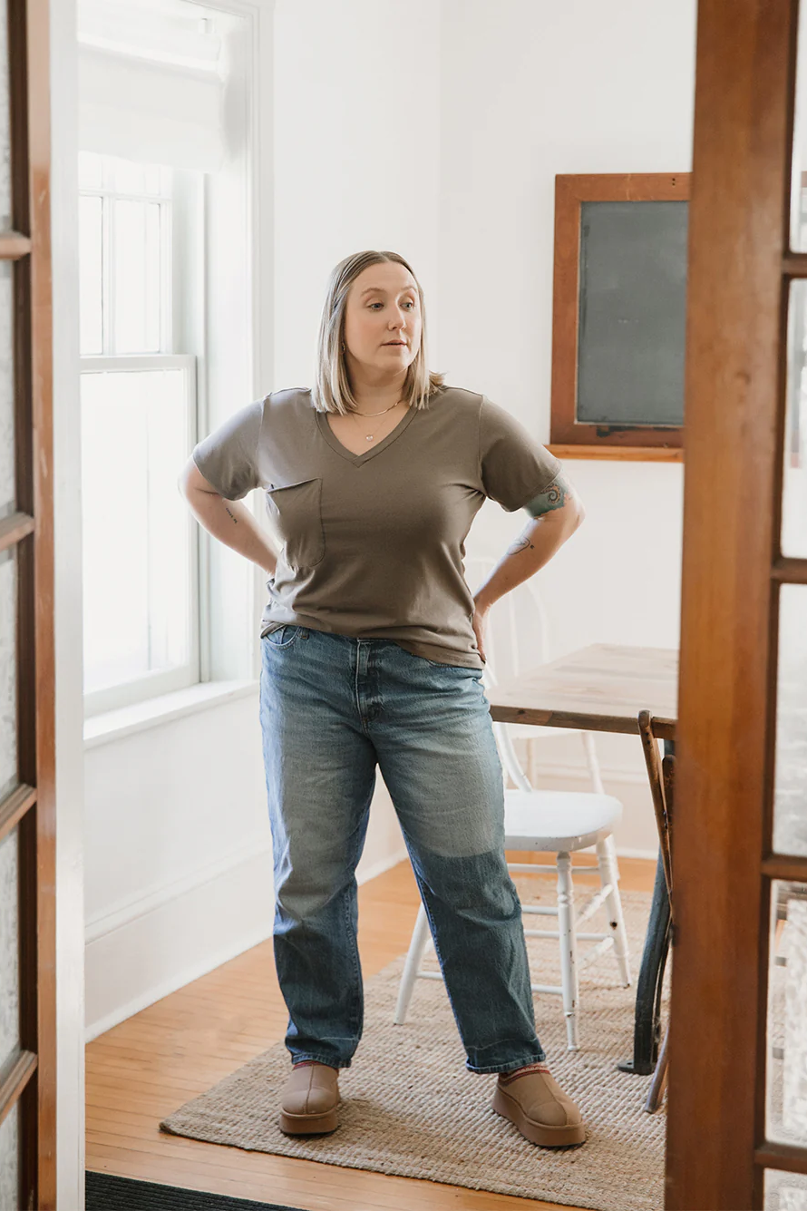 A woman wearing the Wave Tee by Blondie Apparel in Clay with a pair of jeans, standing in front of a table and chairs 