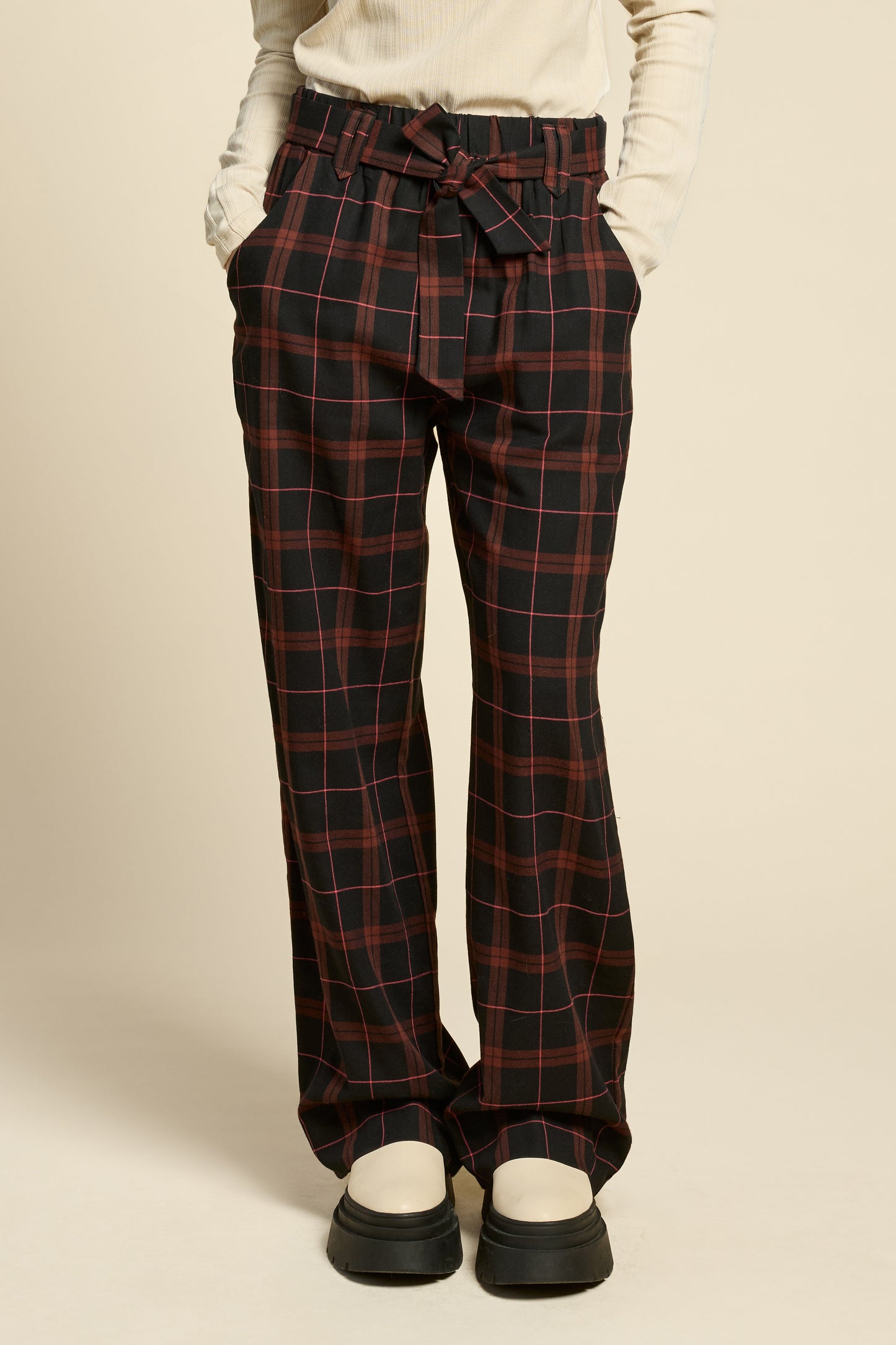 Waist down view of a woman wearing the West Coast Pants by Cokluch in Burgundy Plaid, featuring flared legs, an attached belt with a D-ring closure, and pointed belt loops. She is wearing them with a beige top and standing in front of a beige background.