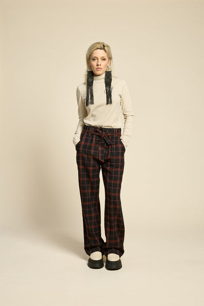 Back view of a woman wearing the West Coast Pants by Cokluch in Burgundy Plaid, featuring flared legs, an attached belt with a D-ring closure, and pointed belt loops. She is wearing them with a beige top and standing in front of a beige background.