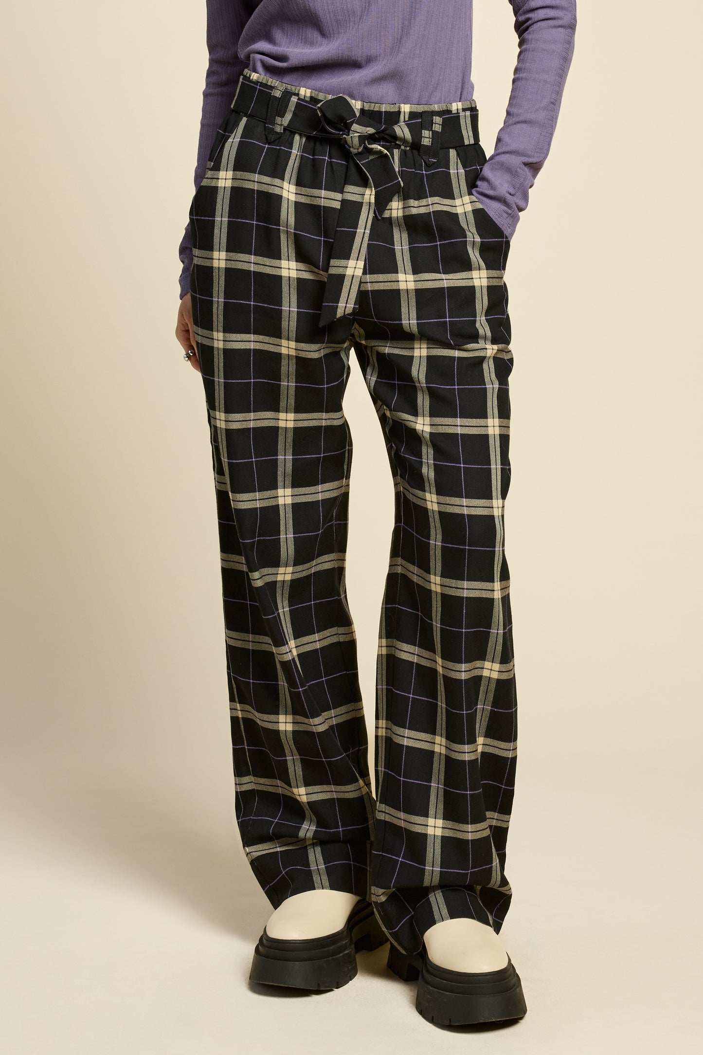 Waist down view of a woman wearing the West Coast Pants by Cokluch in Black Plaid, featuring flared legs, an attached belt with a D-ring closure, and pointed belt loops. She is wearing them with a purple top and standing in front of a beige background.
