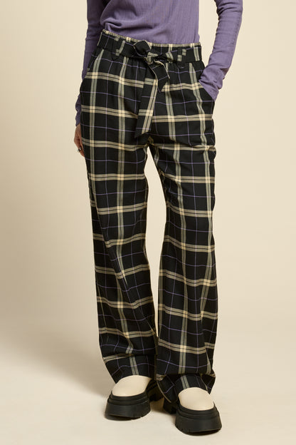 Waist down view of a woman wearing the West Coast Pants by Cokluch in Black Plaid, featuring flared legs, an attached belt with a D-ring closure, and pointed belt loops. She is wearing them with a purple top and standing in front of a beige background.