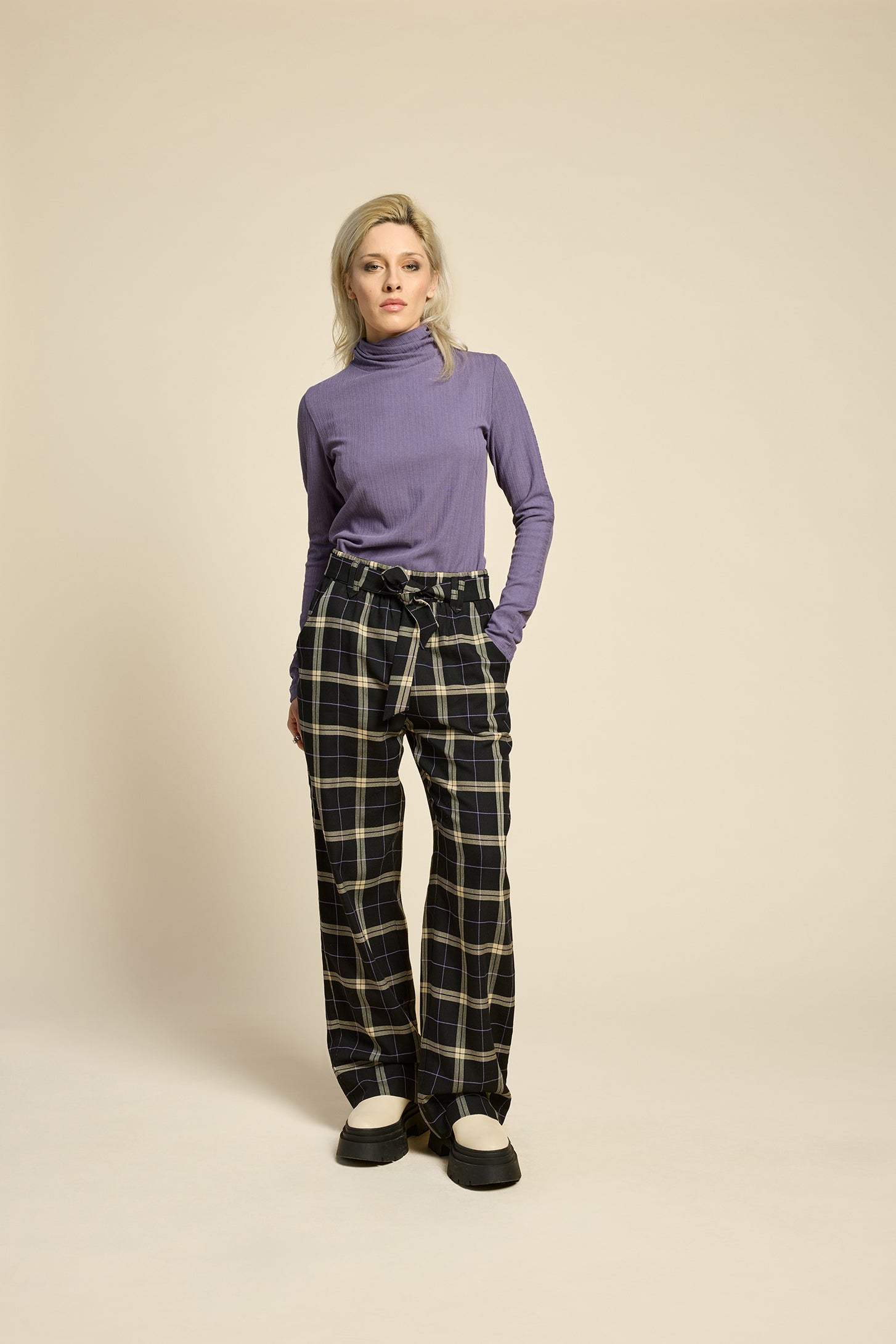 Back view of a woman wearing the West Coast Pants by Cokluch in Black Plaid, featuring flared legs, an attached belt with a D-ring closure, and pointed belt loops. She is wearing them with a purple top and standing in front of a beige background.
