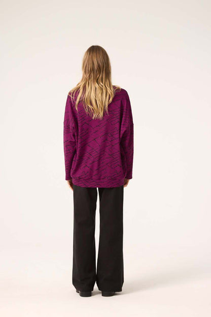 Back view of a woman wearing the Woodland Cardigan by Cokluch in Magenta, with dark pants, standing in front of a white background. 