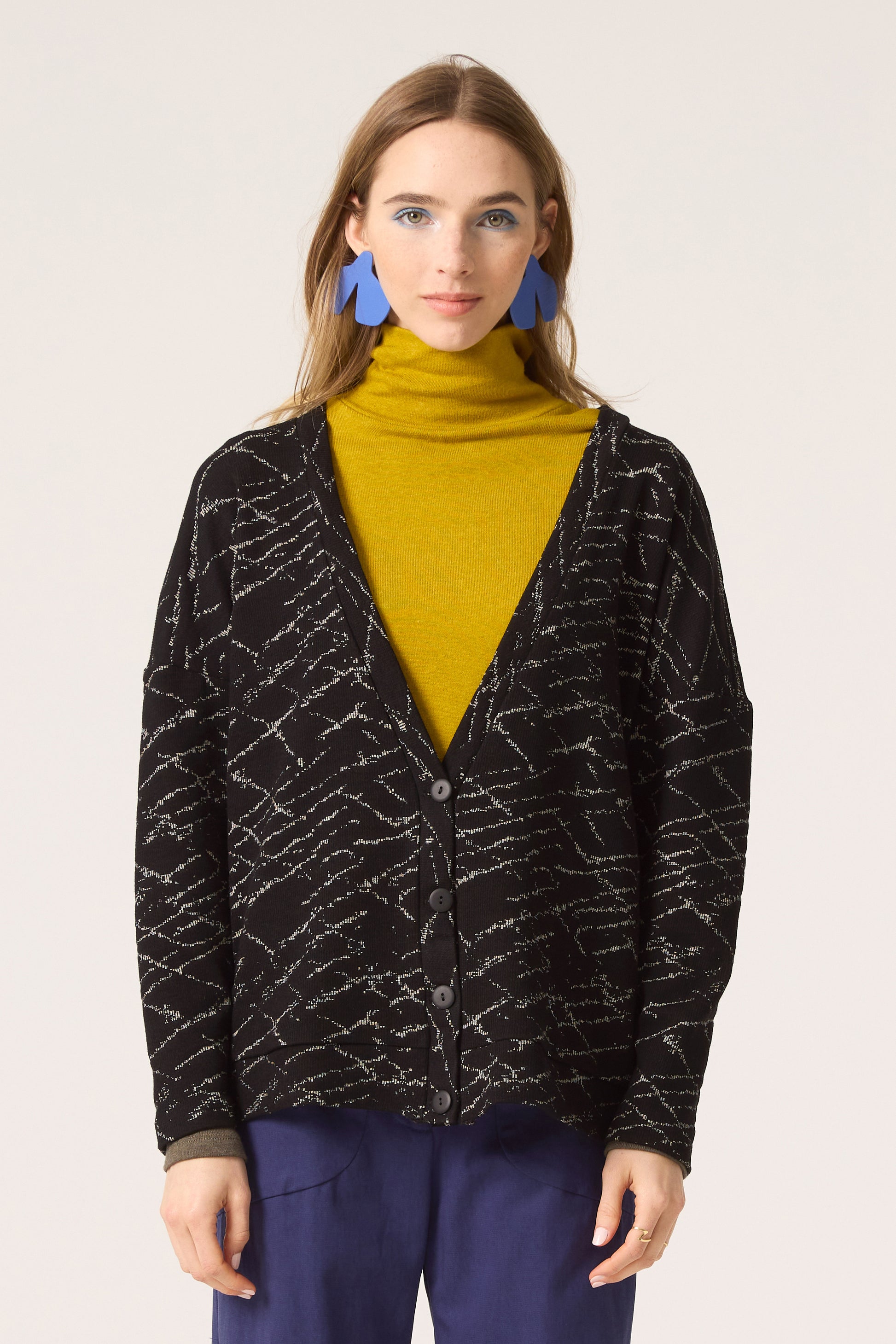 Semi close-up of a woman wearing the Woodland Cardigan by Cokluch in Black, with a yellow top and blue pants, standing in front of a white background. 