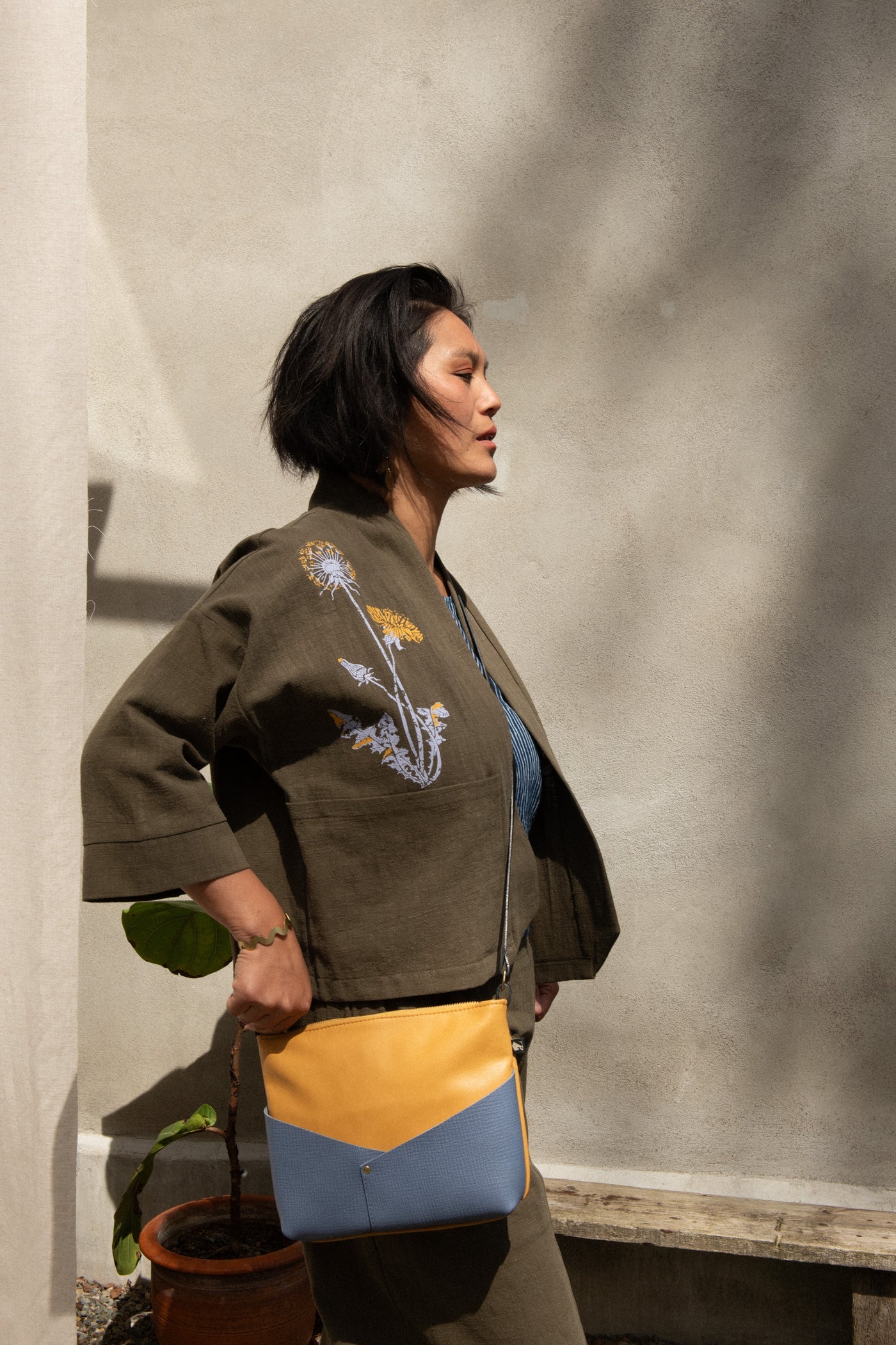 Side view of a woman wearing the Magnolia Screen-printed Jacket from Kazak in Olive with Dandelions, a short kimono style jacket with hand-screen printed dandelions, open front, 3/4-length sleeves, and big patch pockets. She is wearing it with matching pants and a blue and yellow purse and standing outside by a grey wall.