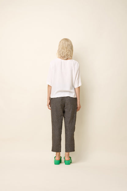 Back view of a woman wearing the Yasmina Pants from Cokluch in Black Utopia, made from checkered linen and cotton with a high elastic waist with a ring belt, and tapered, ankle-length legs. She is wearing them with a white top and standing in front of a beige background. 