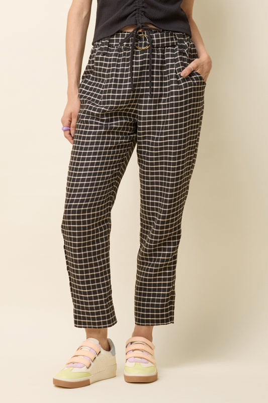 Waist-down view of a woman wearing the Yasmina Pants from Cokluch in Black Utopia, made from checkered linen and cotton with a high elastic waist with a ring belt, and tapered, ankle-length legs. She is wearing them with a black top and standing in front of a white background. 