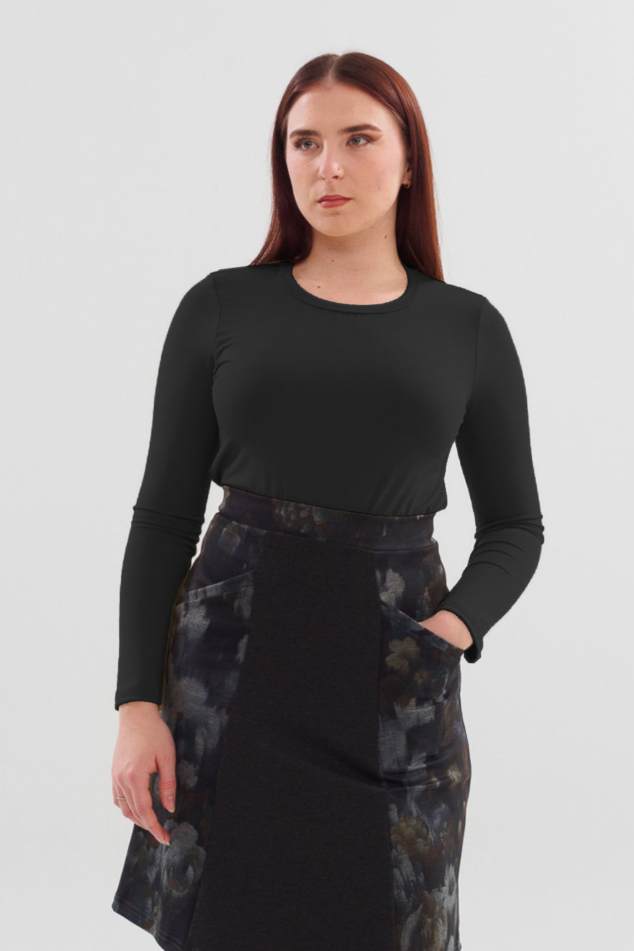 A semi close-up of a woman wearing the A24 Sweater by Slak in Black with  a dark skirt, standing in front of a white background 