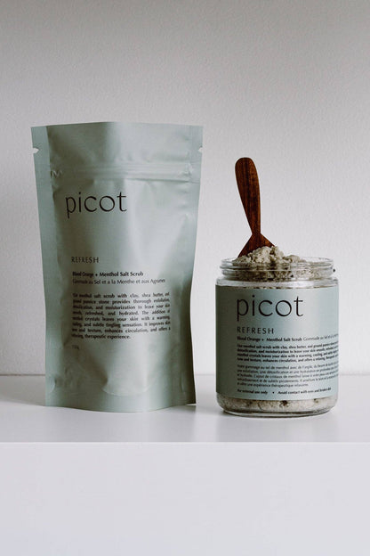 Picot Refresh salt scrub shown with its silver foil pouch and a clear glass jar containing the scrub with a wooden spoon, set against a plain white background.
