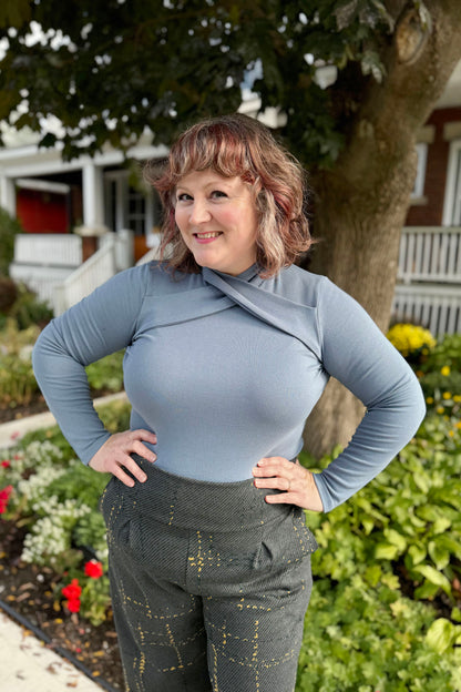 A woman wearing the Irina Top by Julei in Ocean, feauring a crossed collar and long sleeves, with the Jacques Pants by Melow in Anthracite. She is standing outside in front of a garden.  