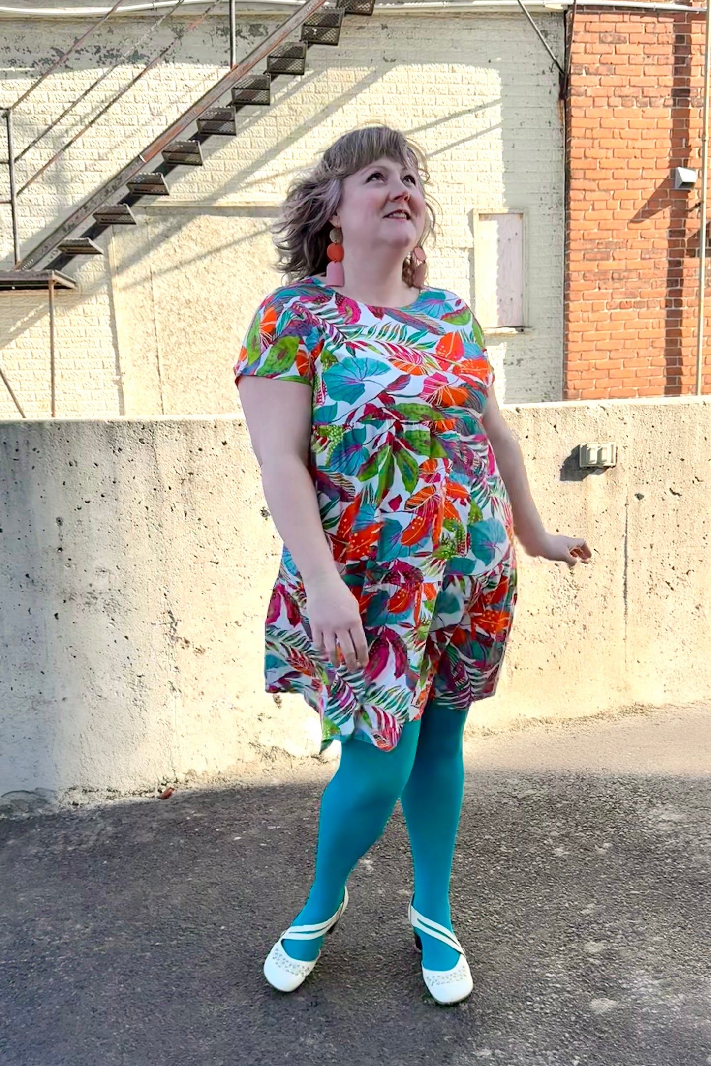 Woman wearing the Grayson Dress by Pure Essence in Peacock Palms, a knee-length baby doll style dress with a tiered skirt and short sleeves. She is standing outside by a brick wall.