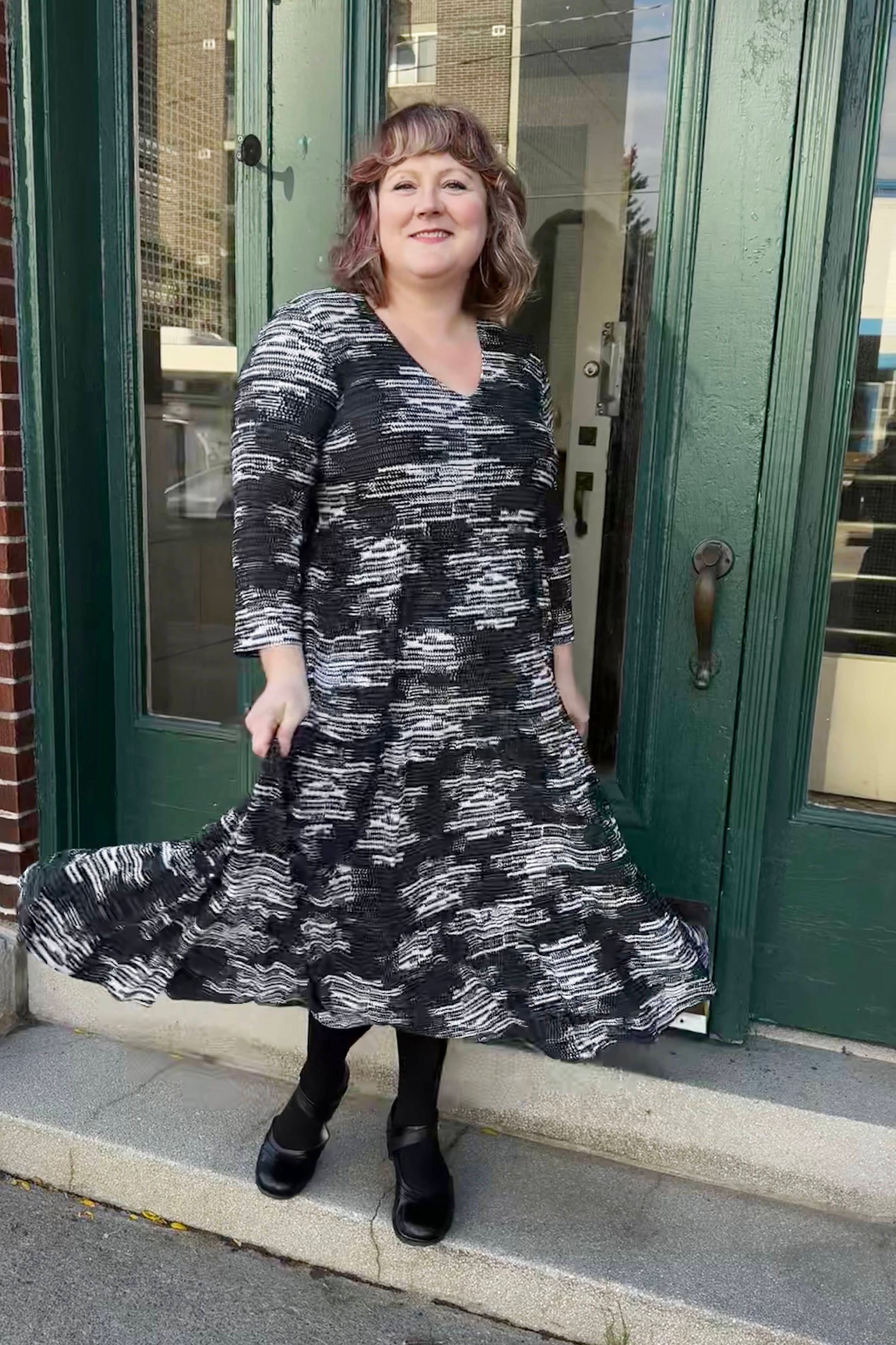 A woman wearing the Duke Dress by Pure Essence in Black/Tan, featuring a V-neck, long sleeves, a gathered waist, and a long tiered skirt, standing in front of a green doorway 