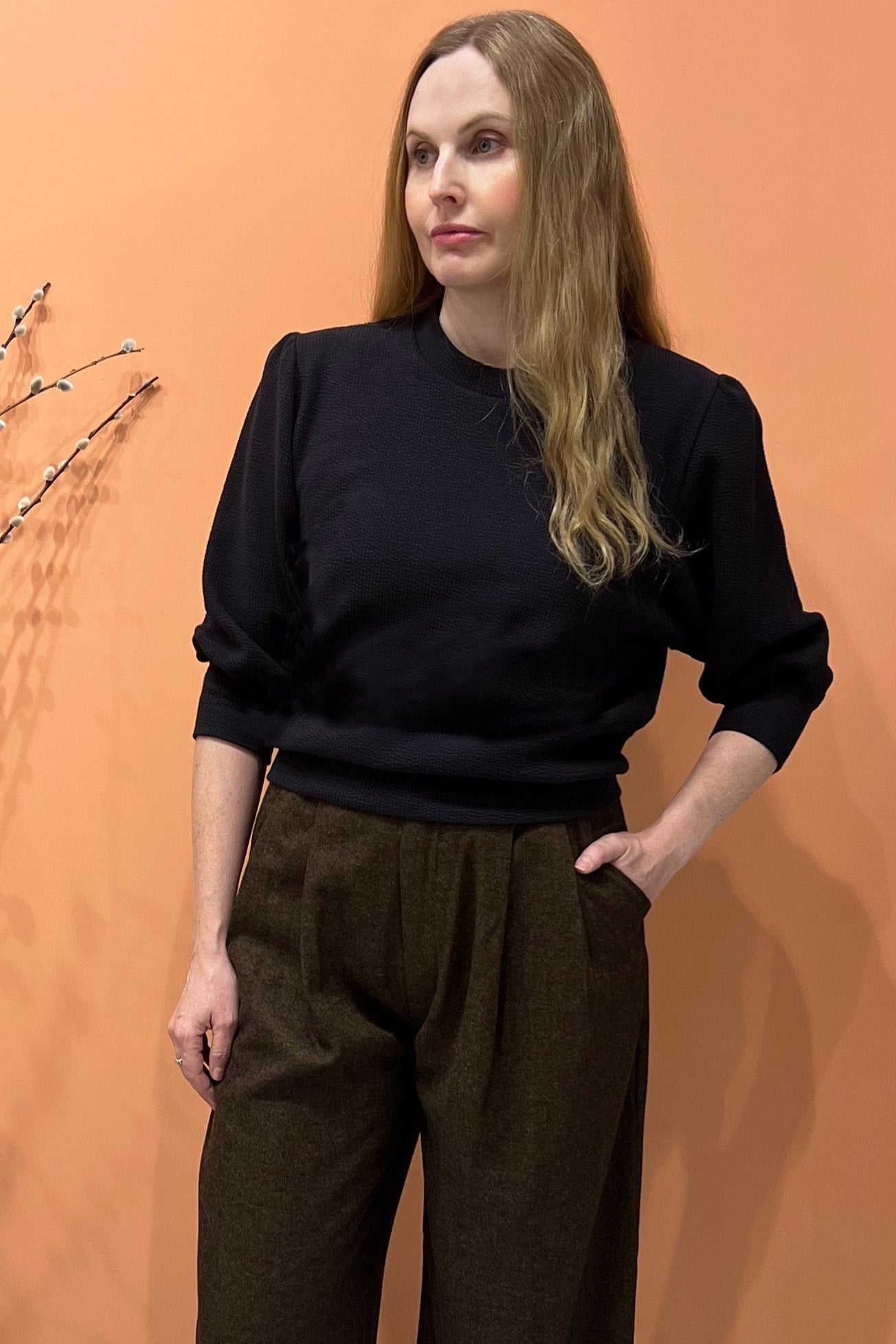 A woman wearing the Bridge Sweater by Allison Wonderland in Grey, featuring elbow length sleeves and a ribbed neckline, cuff, and hem. She's wearing it with the Memory pants in Herringbone and standing in front of an orange wall. 